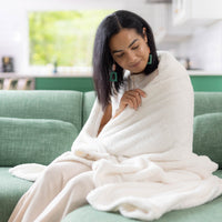 Woman wrapped in a white blanket sitting on a green couch in a bright room.
