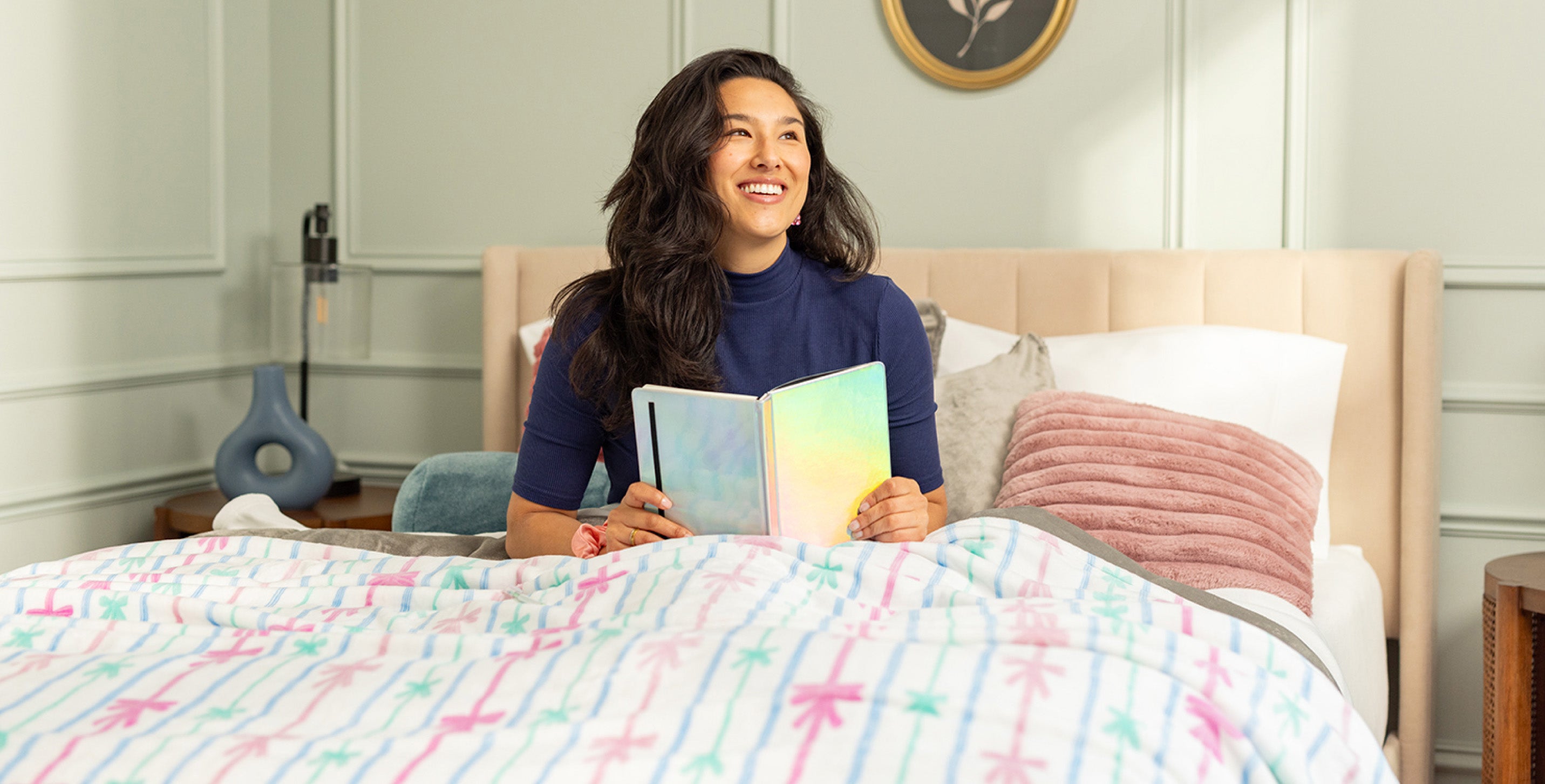 Image of a girl sitting in bed with a silver chrome notebook. The room is pastel themed, featuring a bed blanket with ribbons and bows.