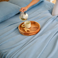 A hand pouring tea onto a wooden tray resting on the All-Season Microfleece Sheet Set in Slate Blue
