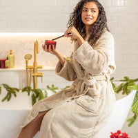 Woman in a beige robe lighting a red candle while in a bathroom setting with Christmas decorations.