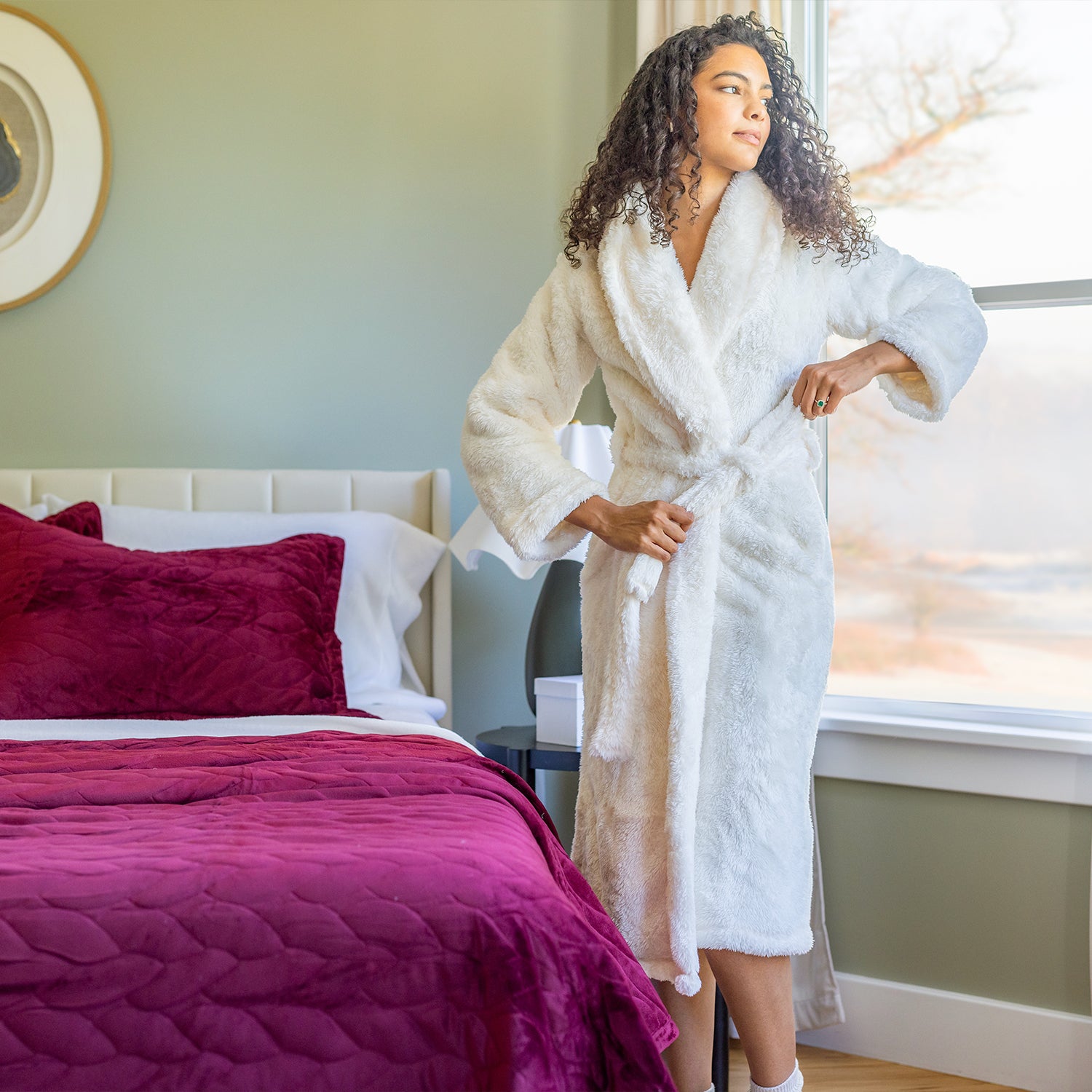 Woman in a white robe standing in a bedroom with purple bedding and a window in the background.