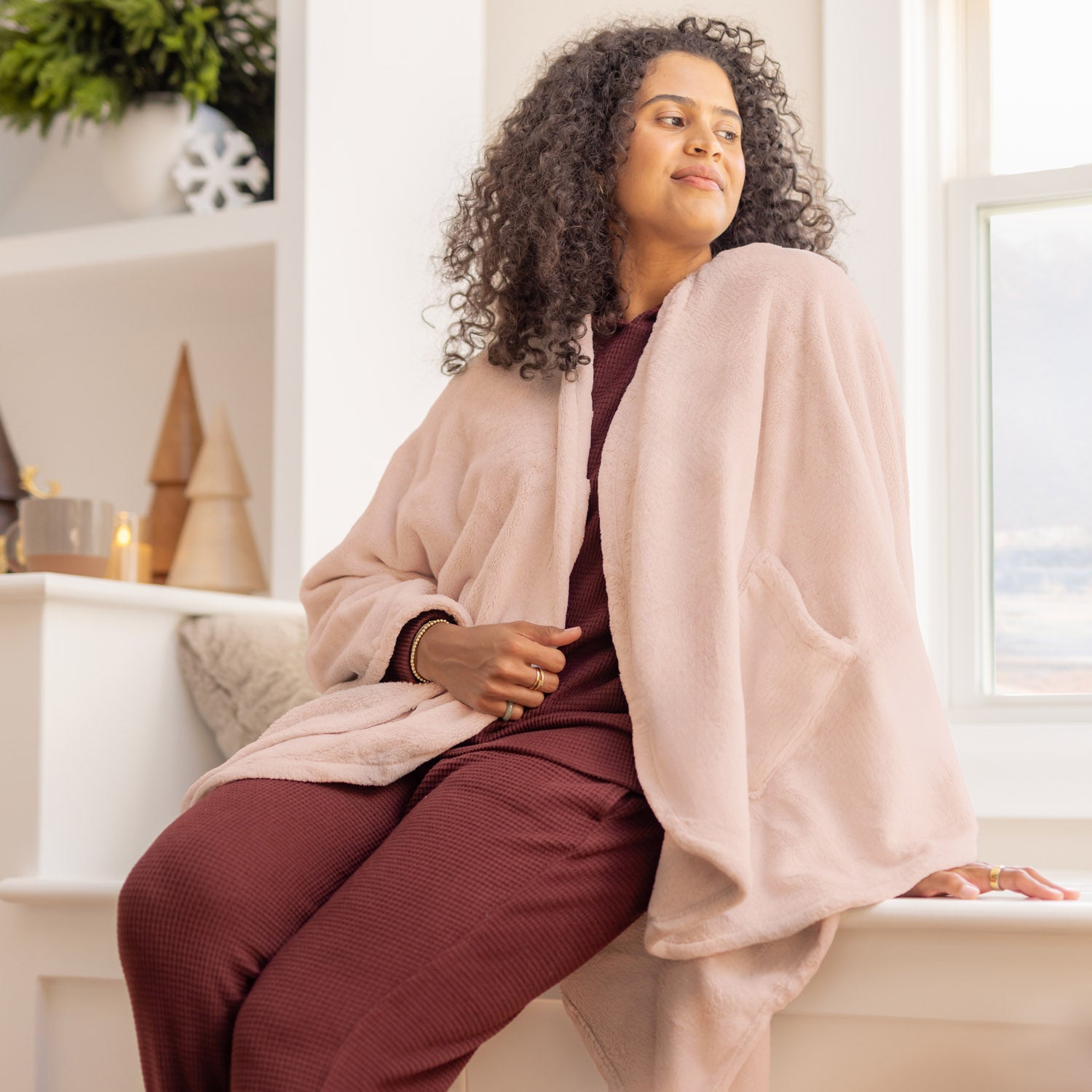 Woman sitting on a couch wearing a pink cape wrap and burgundy pants in a bright room.