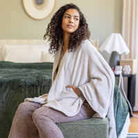 Woman sitting on a bed wearing a white cape wrap in a bedroom setting