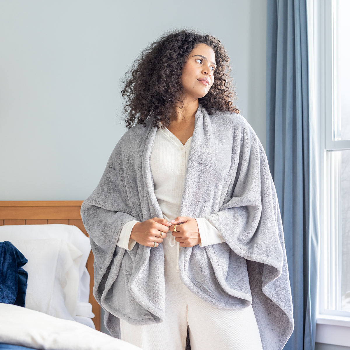 Woman wearing a gray cape wrap draped over her shoulders in a bedroom setting.