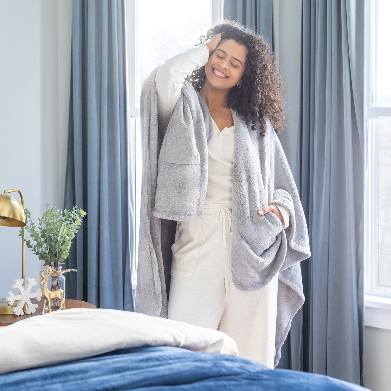 Woman wrapped in a gray cape wrap standing in a bedroom with curtains and a bed.