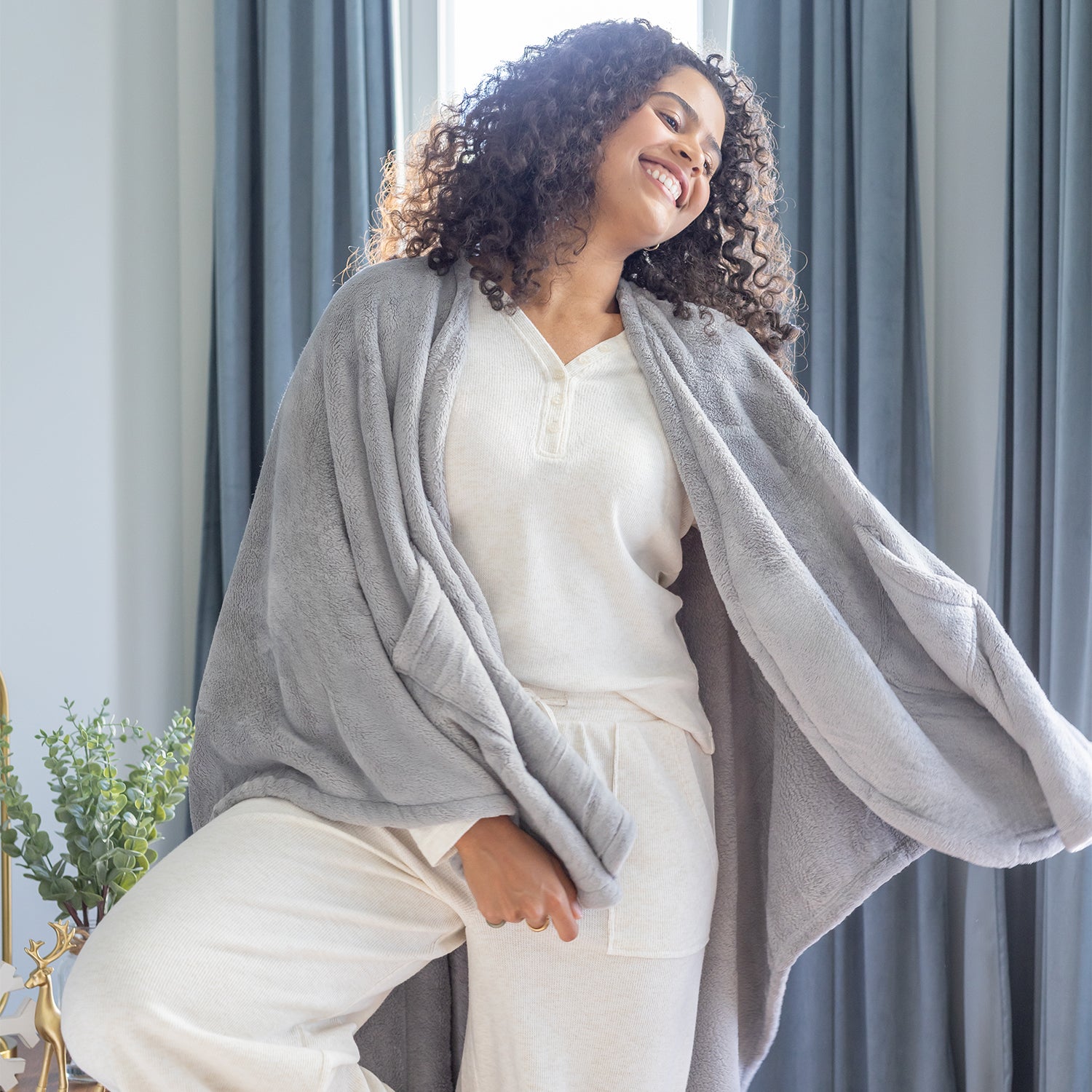 Woman wearing a gray cape wrap over a white outfit in a room with curtains and plants.