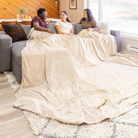 3 people sitting on a grey sofa sharing a beige velvety plush family blanket while smiling.