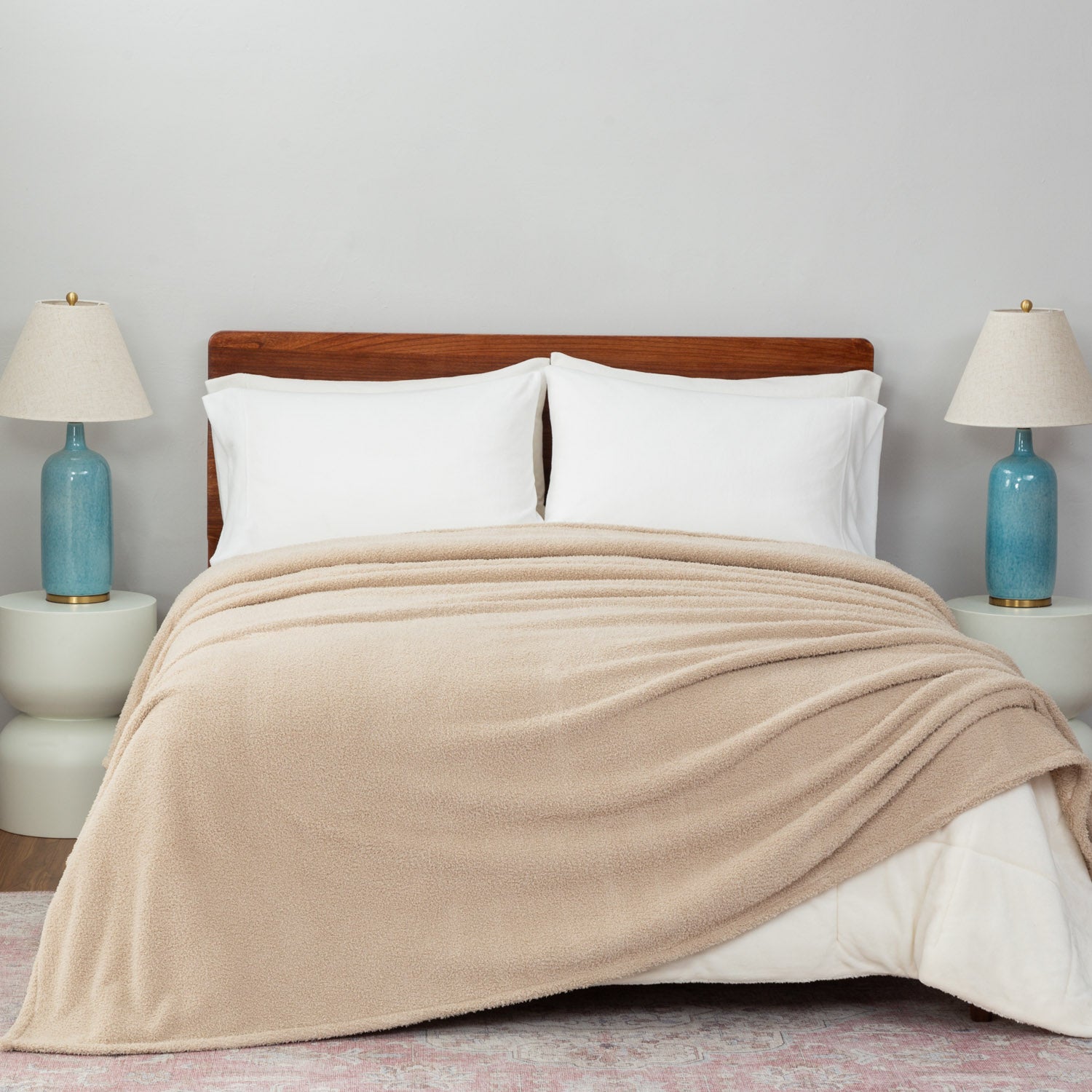 Beige blanket draped over a bed with white pillows and two lamps on nightstands.