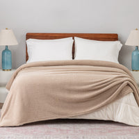 Beige blanket draped over a bed with white pillows and wooden headboard, against a light gray wall.