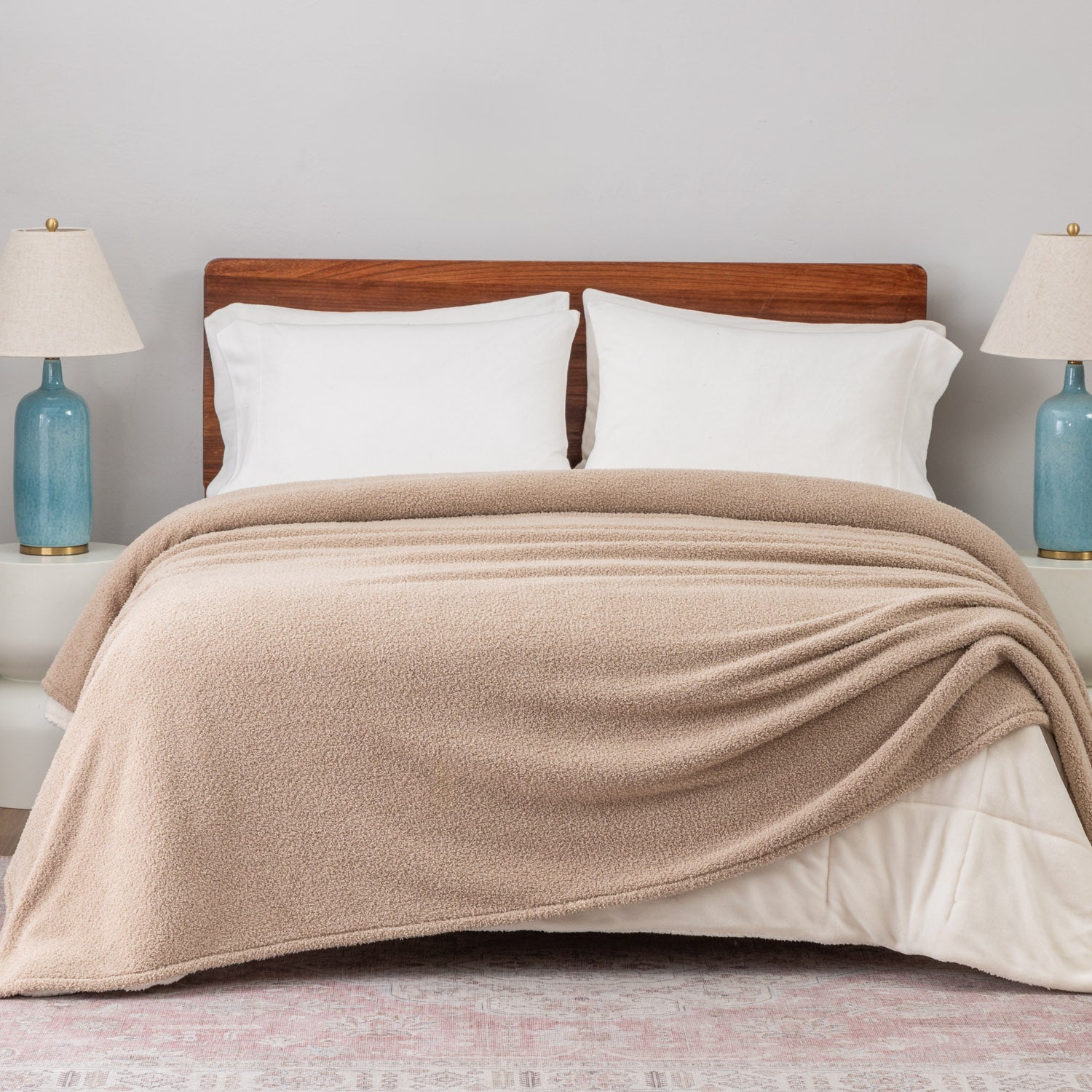 Beige blanket draped over a bed with white pillows and wooden headboard, against a light gray wall.