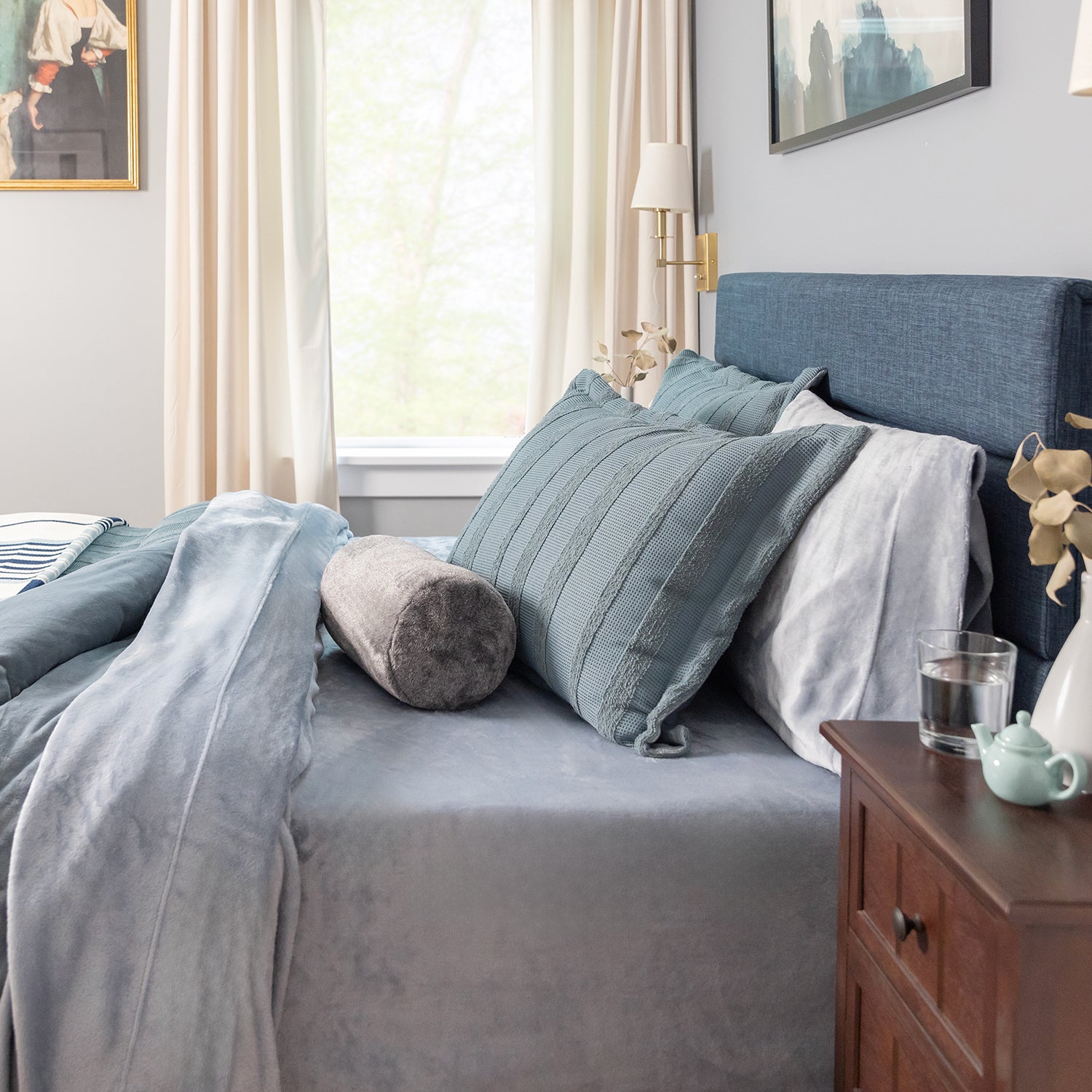 Cozy bedroom with a bed featuring striped bedding and pillows, a wooden nightstand with a teapot and glass, and a window with curtains.