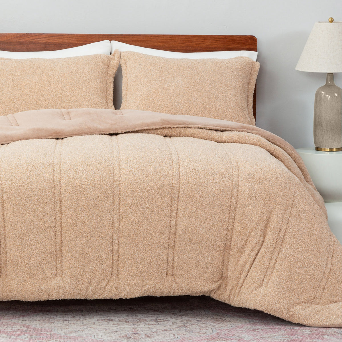Beige textured comforter on a bed with pillows and a lamp in the background.