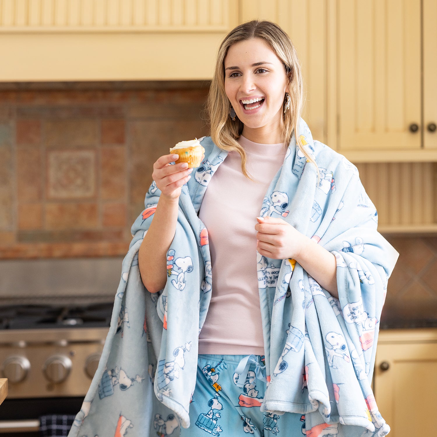 A woman wrapped in a blue peanuts printed throw while holding a cupcake.