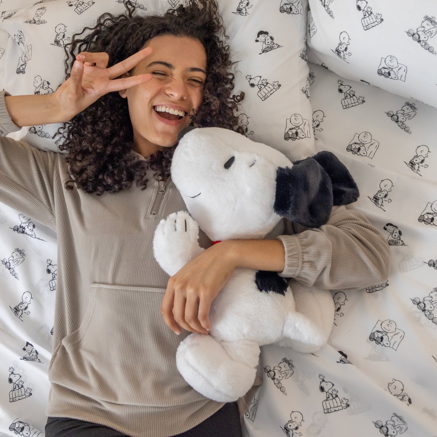 Person holding a Snoopy plush toy on a bed with Snoopy and Woodstock bedding.