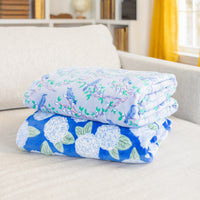Two folded velvetloft throws with printed designs, one with a blue floral pattern and another with a white and blue animal pattern, draped over the arm of a sofa.