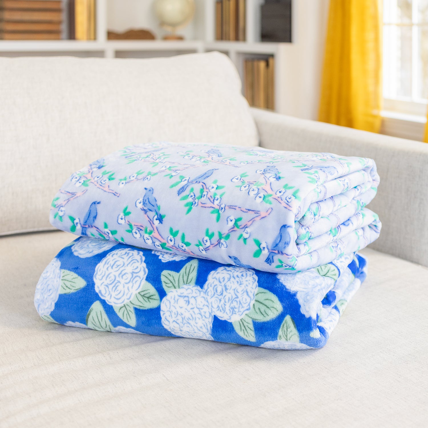 Two folded velvetloft throws with printed designs, one with a blue floral pattern and another with a white and blue animal pattern, draped over the arm of a sofa.