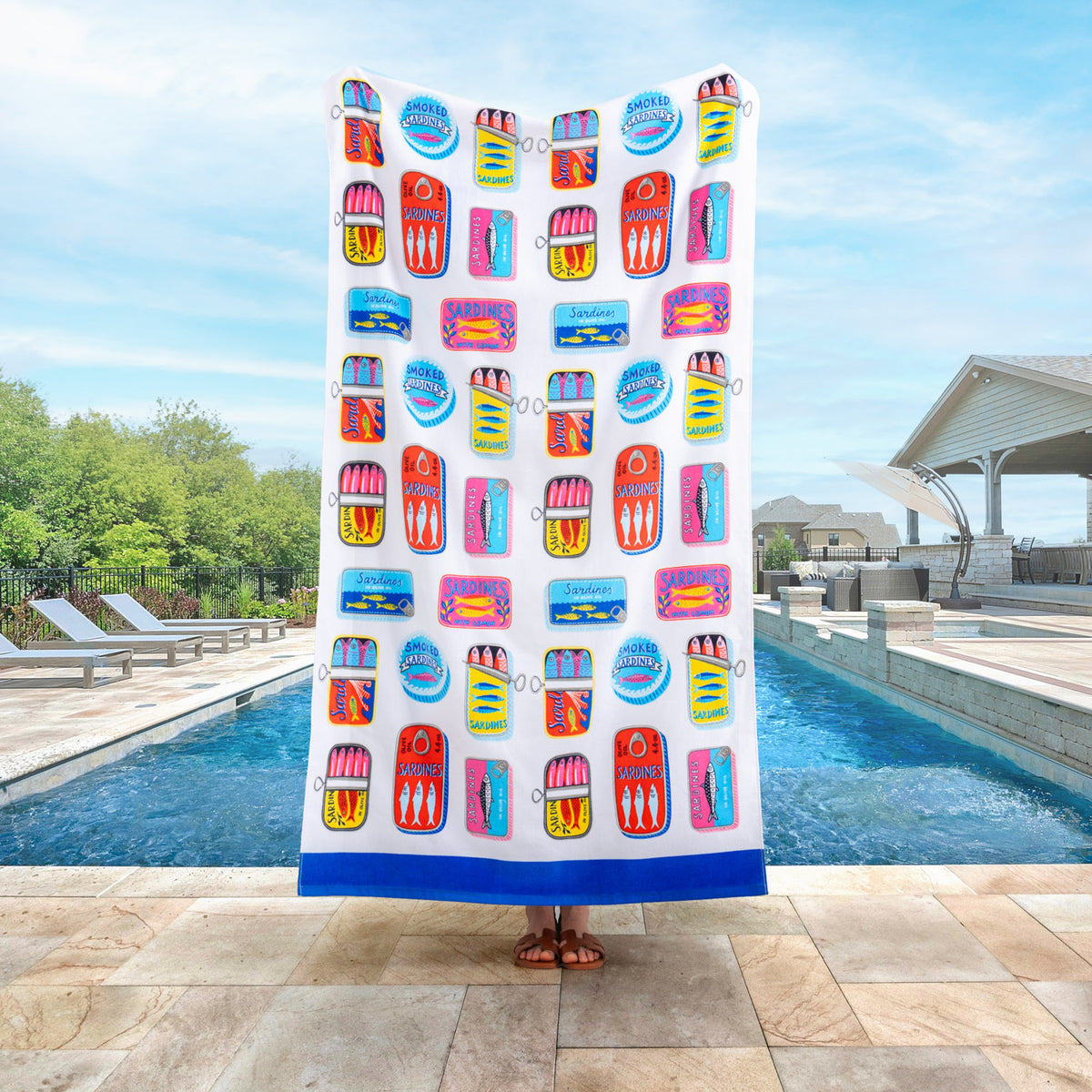 Bex Morley Sardine Tin beach towel held up by a person standing by a pool.