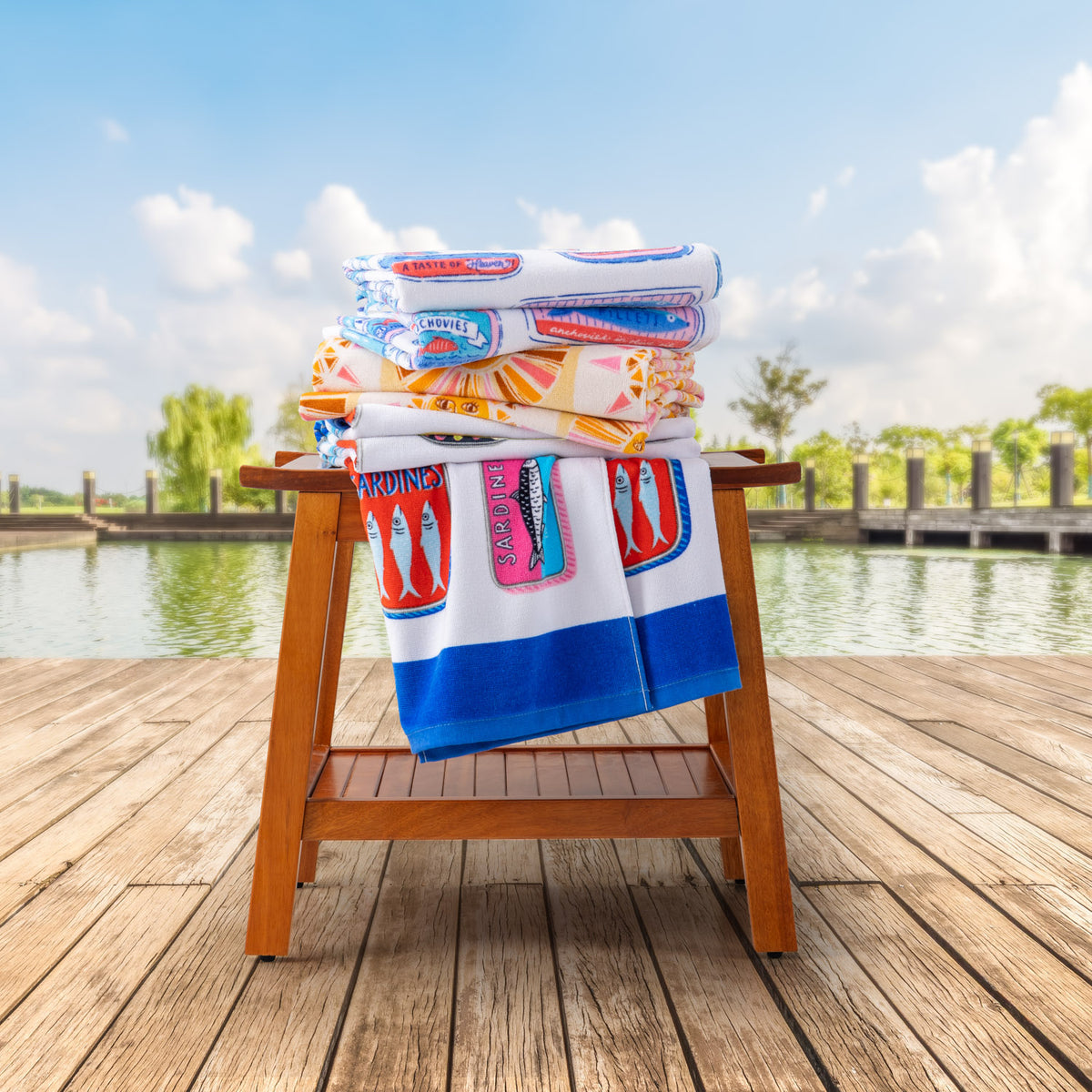 A stack of 3 printed beach towels stacked on top of a wooden chair by a pond.