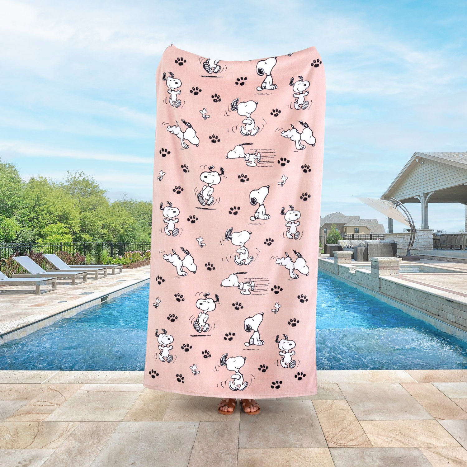 A person holding up a pink Peanuts beach towel while standing by a pool.