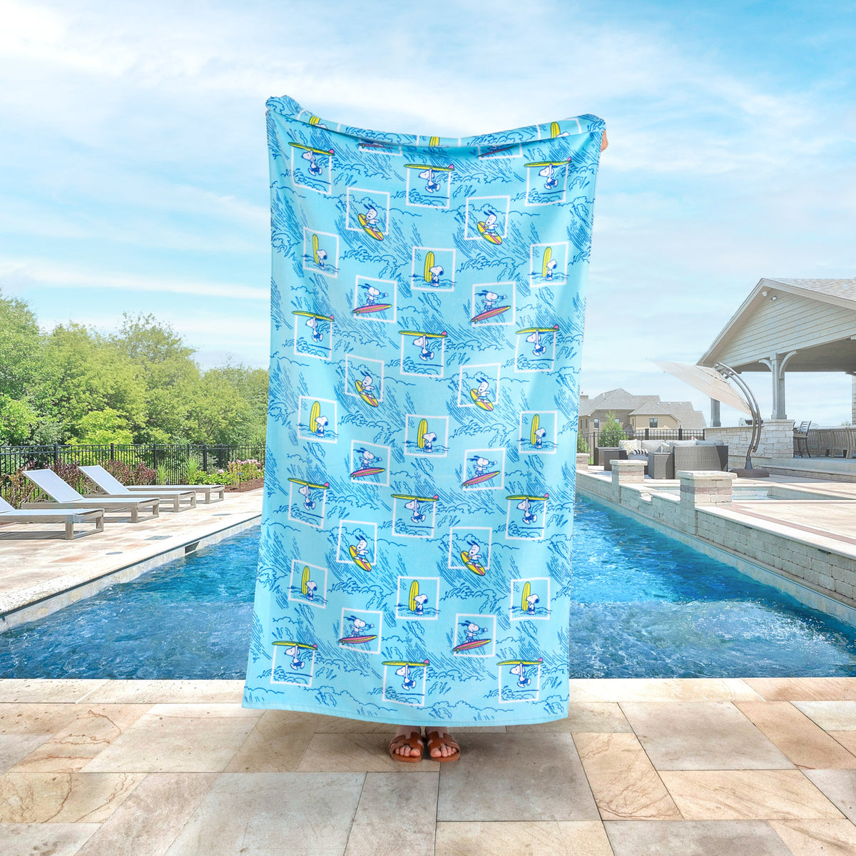 A blue peanuts beach towel held up by a person standing by a pool.