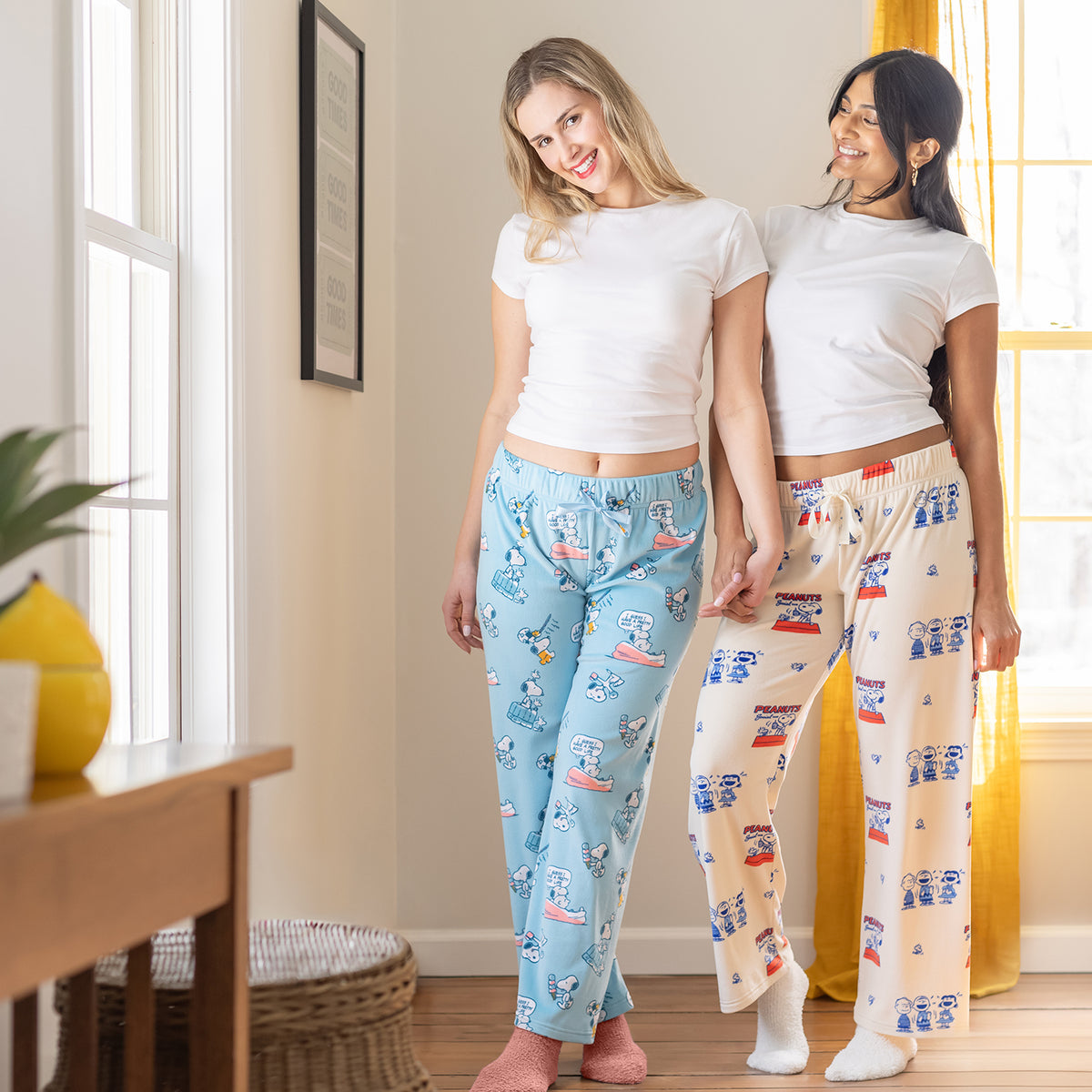 Two woman wearing Peanuts printed lounge pants while smiling and holding hands.