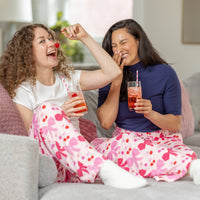 Two women in pajama pants sitting on a couch, enjoying drinks and a cherry.