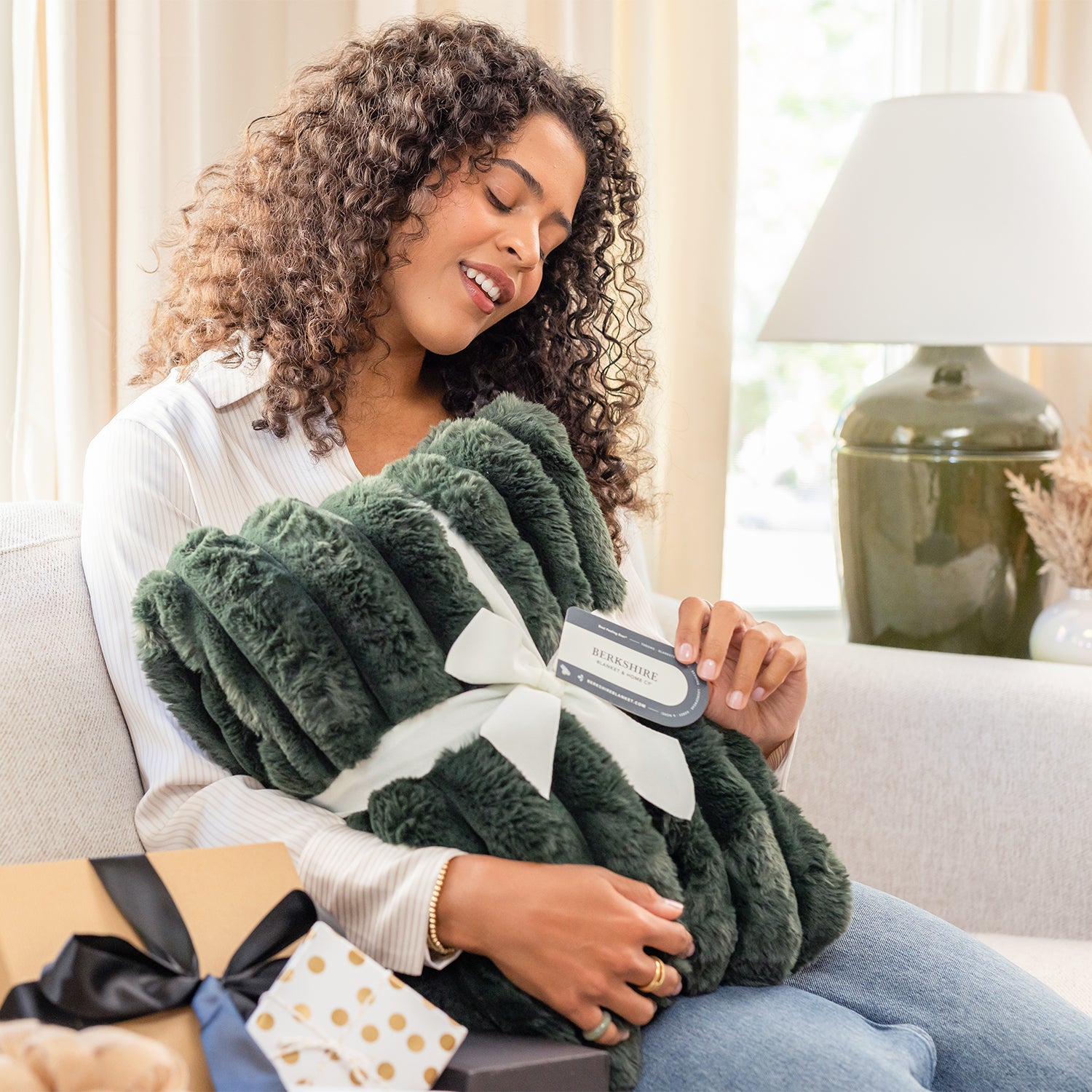 Woman holding a green fluffy blanket with a gift card, sitting on a couch in a cozy living room.