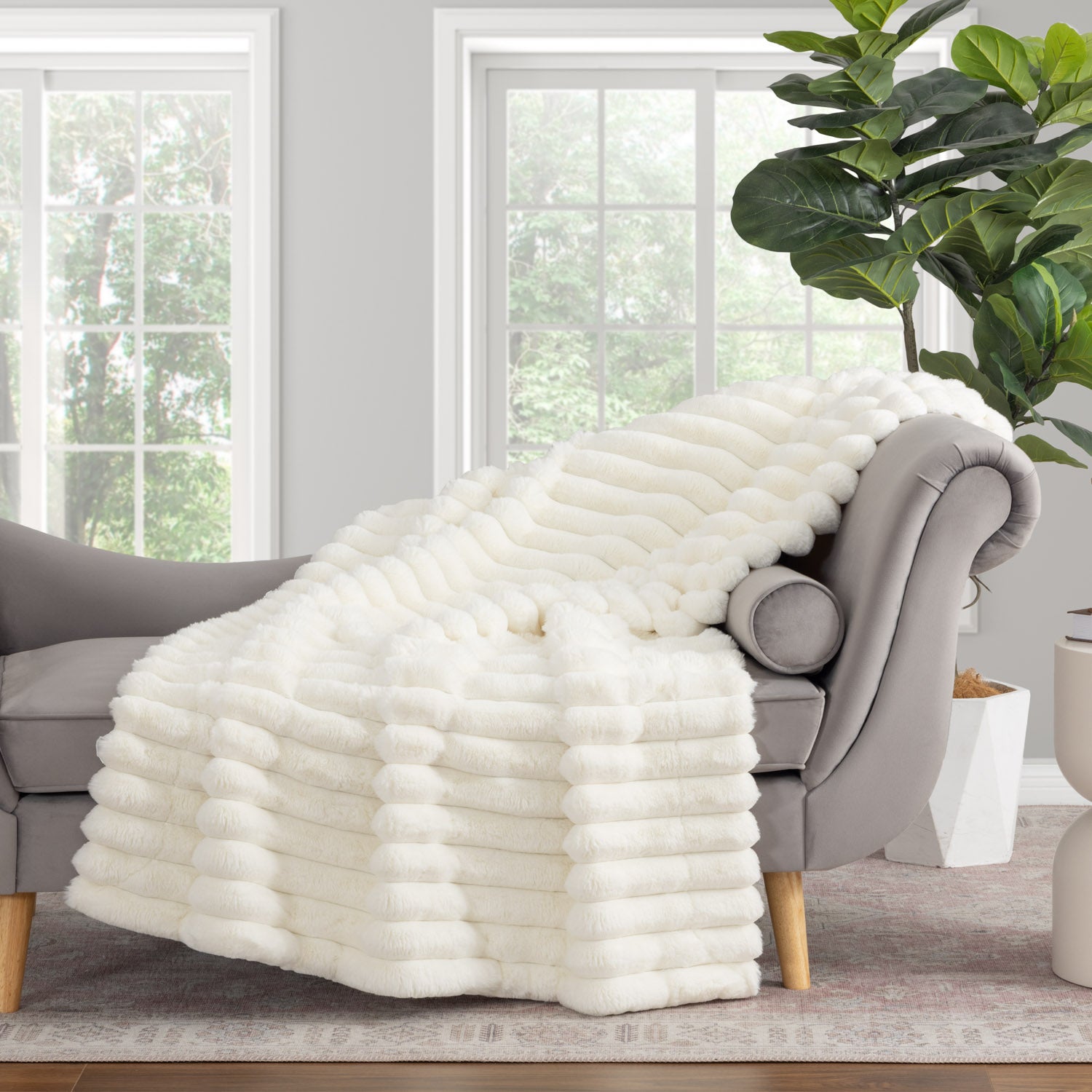 White fluffy blanket draped over a gray armchair in a living room with large windows and a plant.
