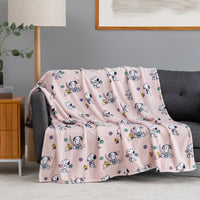 A pink fleece throw blanket with a repeating cartoon Snoopy and Woodstock pattern is draped over a dark gray sofa in a living room setting.