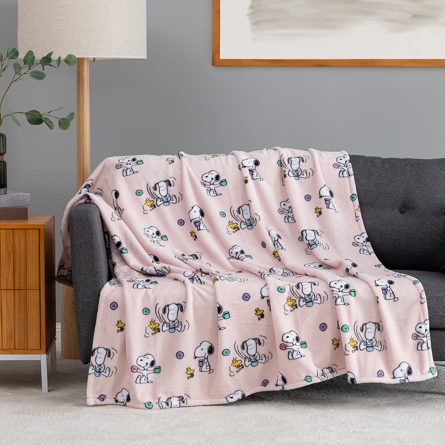 A pink fleece throw blanket with a repeating cartoon Snoopy and Woodstock pattern is draped over a dark gray sofa in a living room setting.