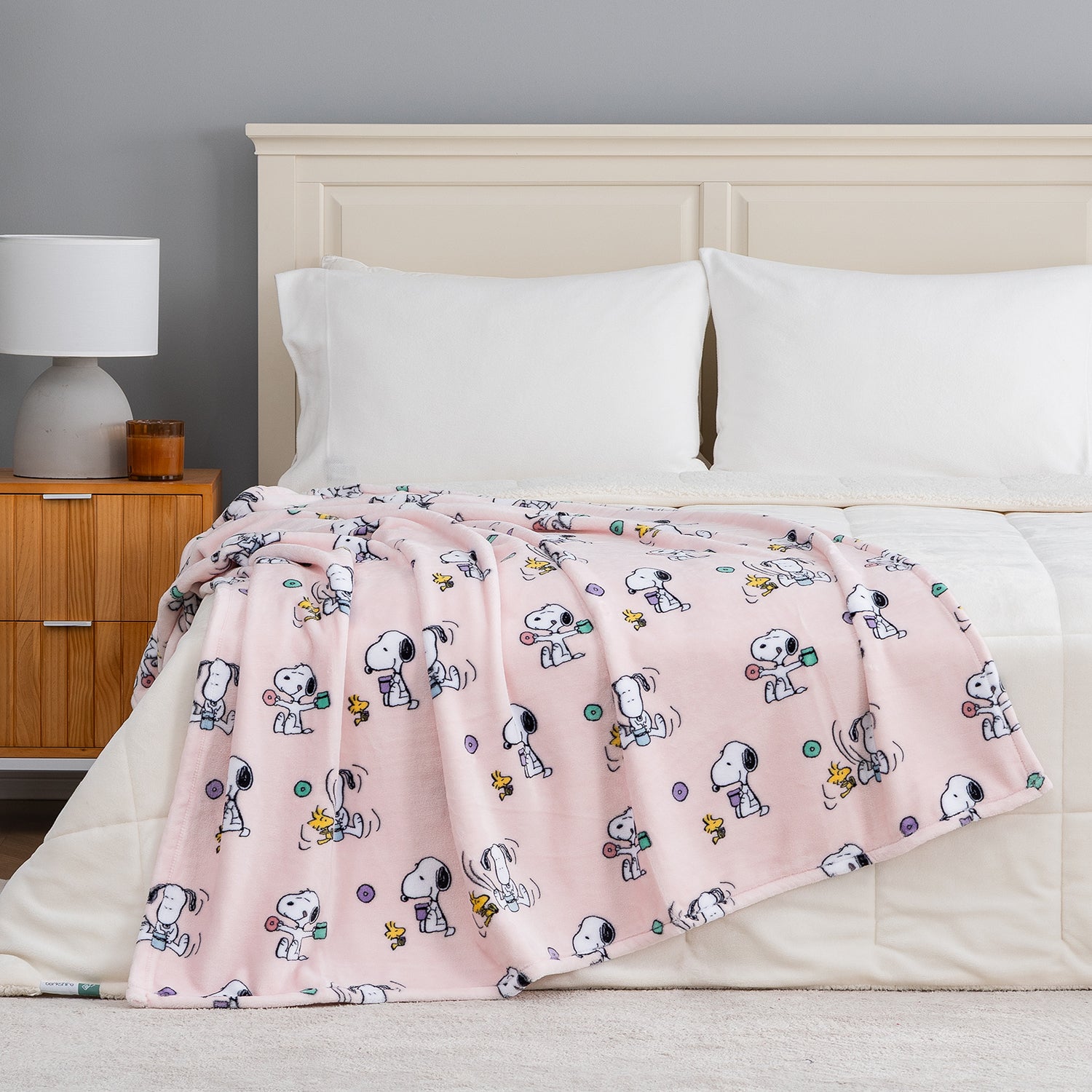 A pink fleece throw blanket featuring a repeating cartoon dog character print, draped over a bed with white bedding in a bedroom setting.