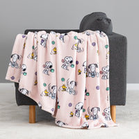 A light pink fleece throw blanket with a cartoon Snoopy and Woodstock print, draped over a dark gray armchair with wooden legs against a neutral gray background.