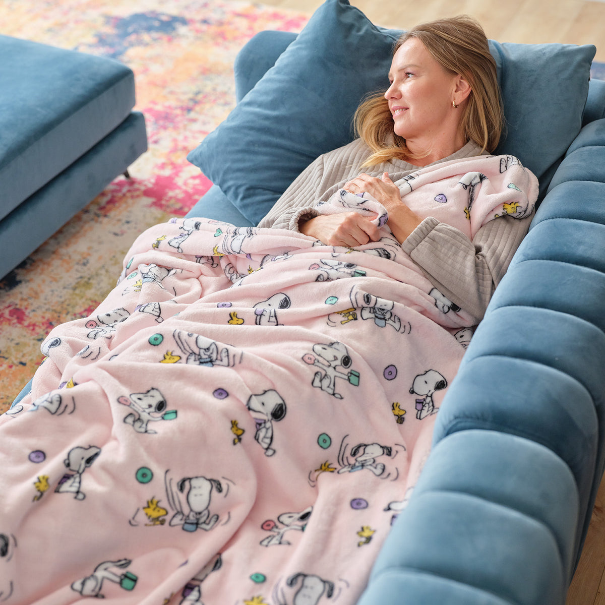 A woman laying on a blue sofa with a pink Peanuts throw blanket draped over her.