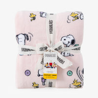 A folded light pink plush blanket with a Peanuts character print, tied with a white ribbon and hangtag. The blanket features Snoopy, Woodstock, and other Peanuts characters on a clean white background.