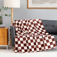 A checkered throw blanket draped over a gray sofa in a living room setting, featuring alternating dark brown and cream squares with small embroidered monogram details.