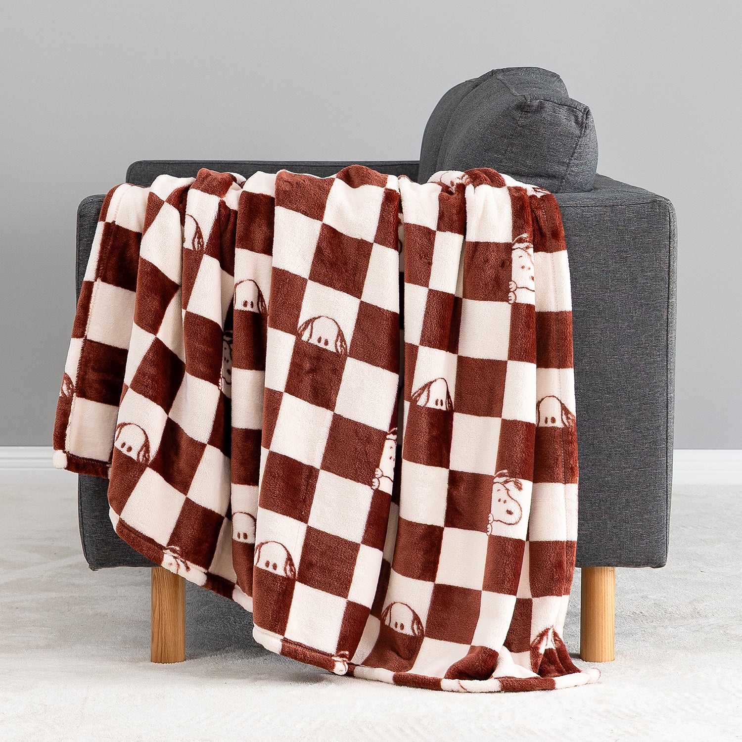 A plush throw blanket with a brown and cream checkerboard pattern and small cartoon dog faces, draped over a dark gray armchair in a studio setting.