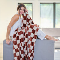 A woman sitting on the edge of a grey sofa holding a Peaunts checkered throw blanket.
