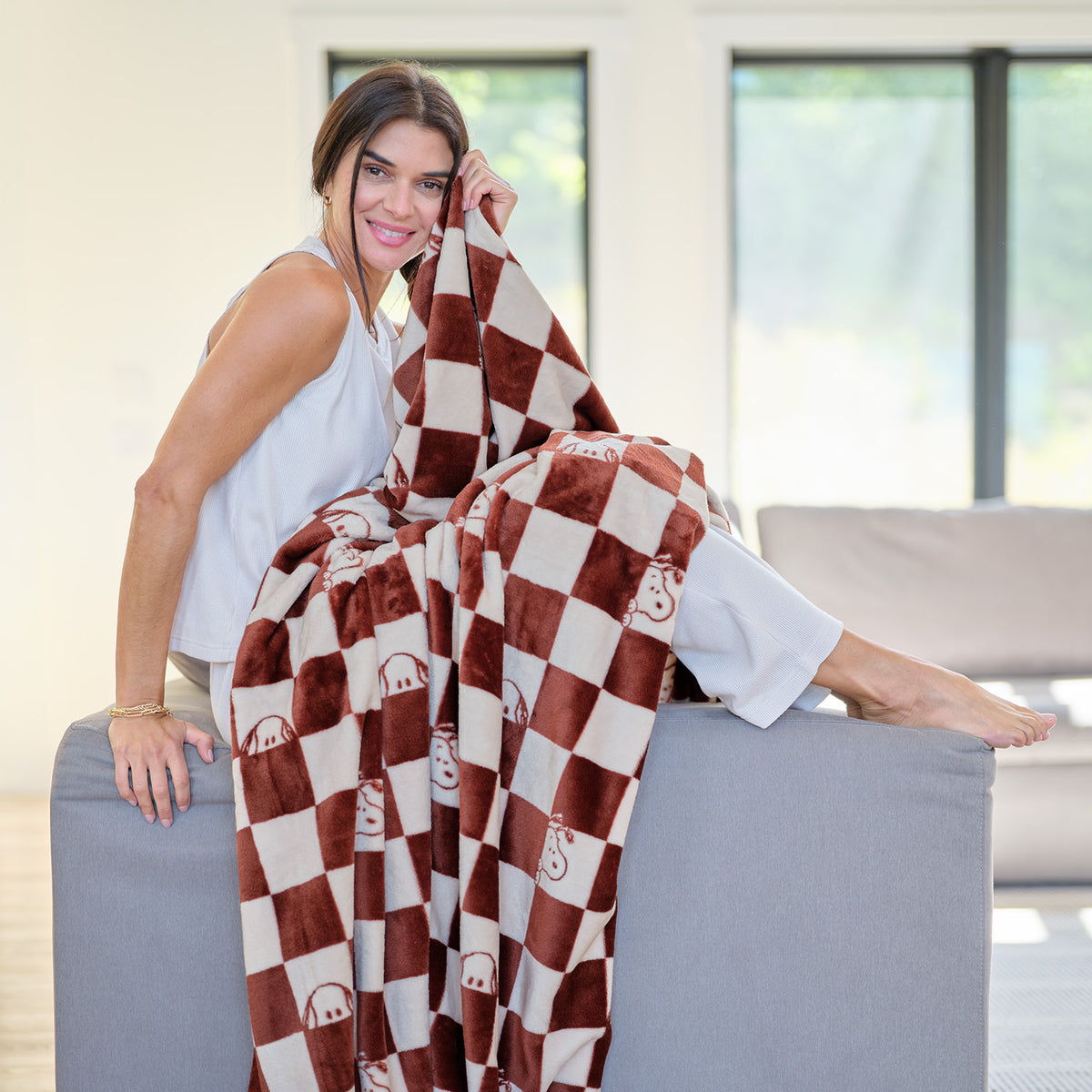 A woman sitting on the edge of a grey sofa holding a Peaunts checkered throw blanket.