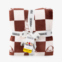 A folded plush throw blanket with a brown and cream checkered pattern, featuring cartoon Peanuts characters. The blanket is wrapped in a white ribbon printed with the 'Peanuts' brand name and tied with a paper hangtag.