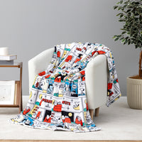 A white throw blanket draped over a white armchair in a styled room, featuring a colorful cartoon comic strip print with Snoopy and other Peanuts characters.