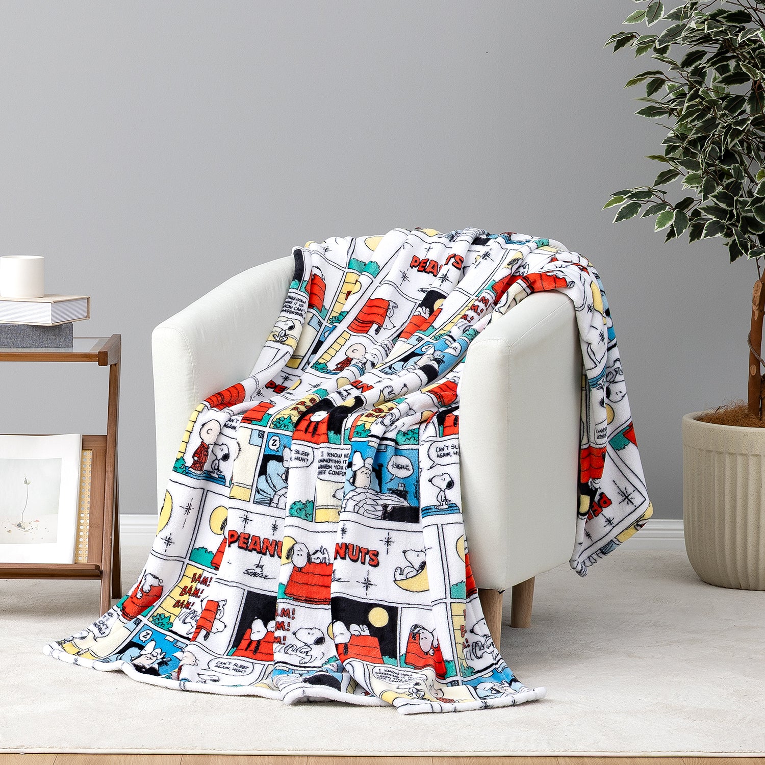A white throw blanket draped over a white armchair in a styled room, featuring a colorful cartoon comic strip print with Snoopy and other Peanuts characters.
