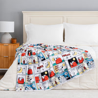 A bed with white bedding and pillows, draped with a colorful Snoopy and Charlie Brown comic strip patterned blanket. The scene is set in a bedroom with a wooden nightstand, a gray wall, and a white lamp.