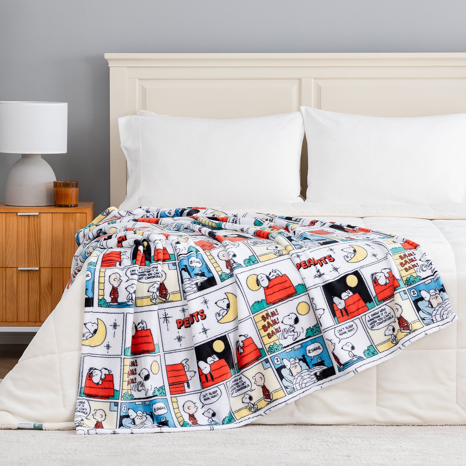 A bed with white bedding and pillows, draped with a colorful Snoopy and Charlie Brown comic strip patterned blanket. The scene is set in a bedroom with a wooden nightstand, a gray wall, and a white lamp.