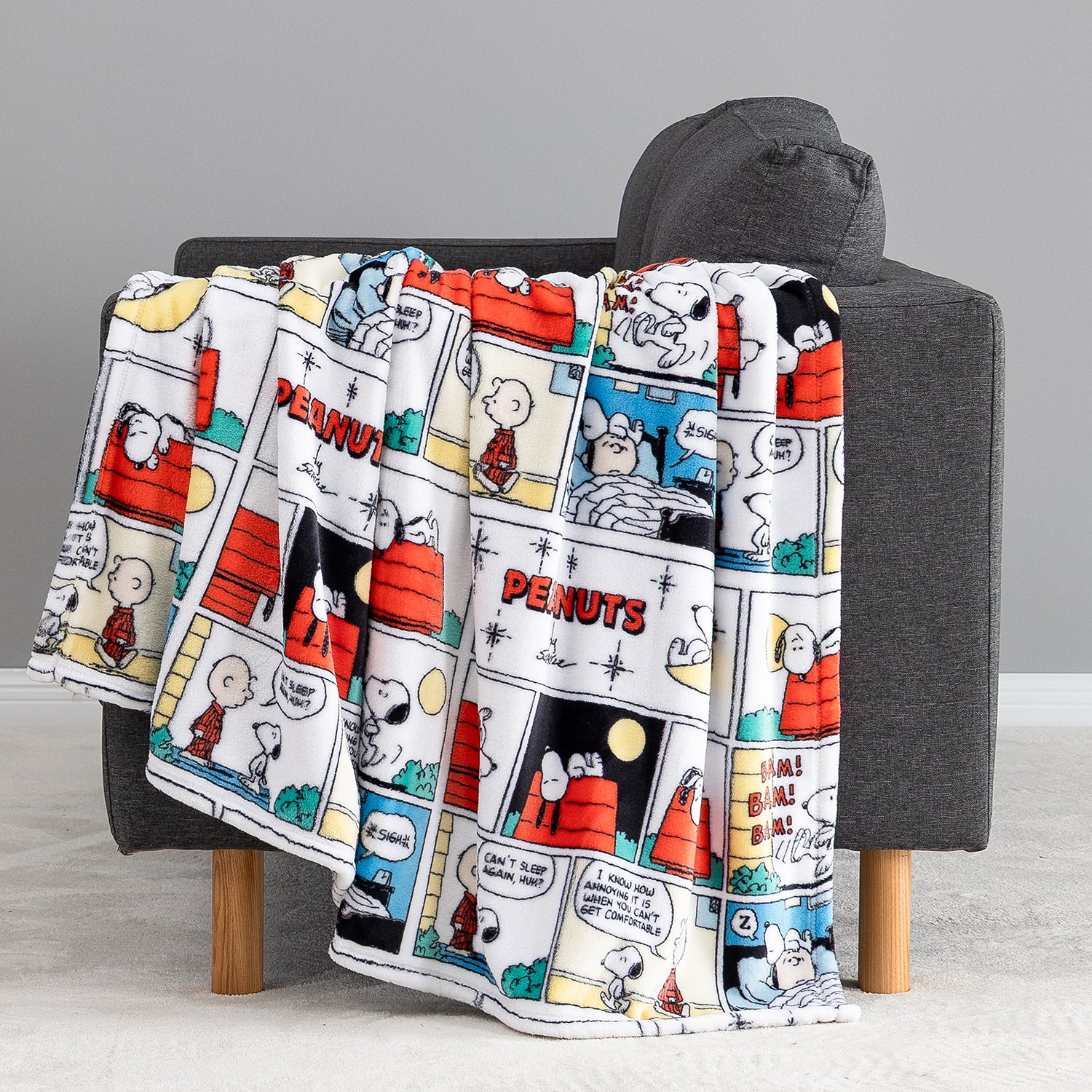 A fleece throw blanket featuring a classic Peanuts comic strip print draped over a dark gray armchair.