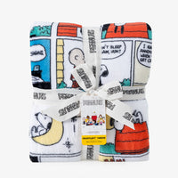 A folded plush throw blanket featuring Peanuts comic strip characters and scenes, tied with a white branded ribbon and a small yellow care tag, on a light gray background.
