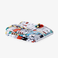A folded blanket featuring a colorful cartoon comic strip print of Snoopy and Woodstock on a white background, set against a plain light gray studio backdrop.
