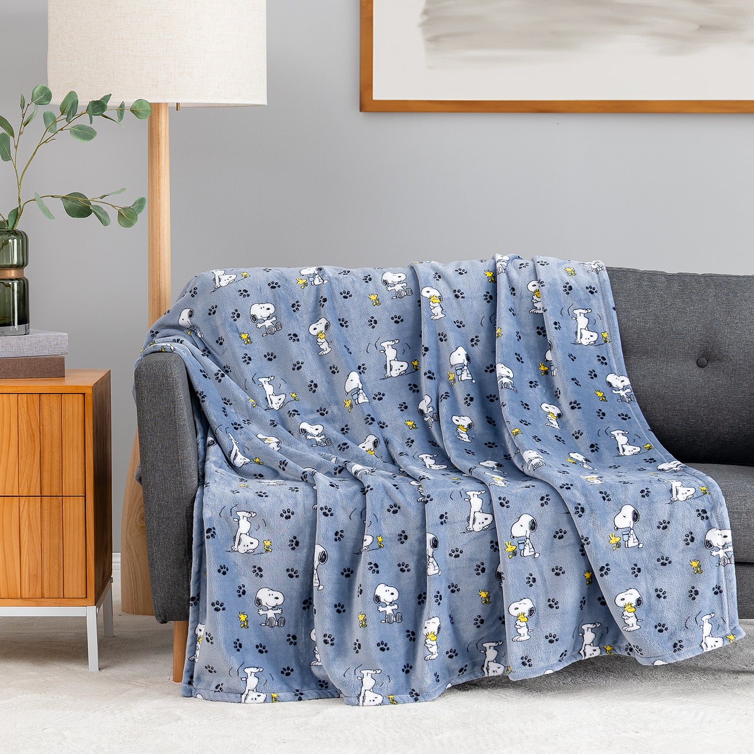 A blue throw blanket with a repeating pattern of white cartoon dogs, black paw prints, and small yellow flowers, draped over a gray sofa in a living room setting.