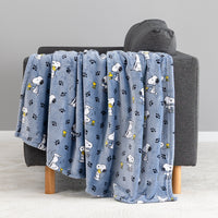 A light blue plush throw blanket with a repeating pattern of Snoopy, Woodstock, and paw prints, draped over a dark gray armchair.