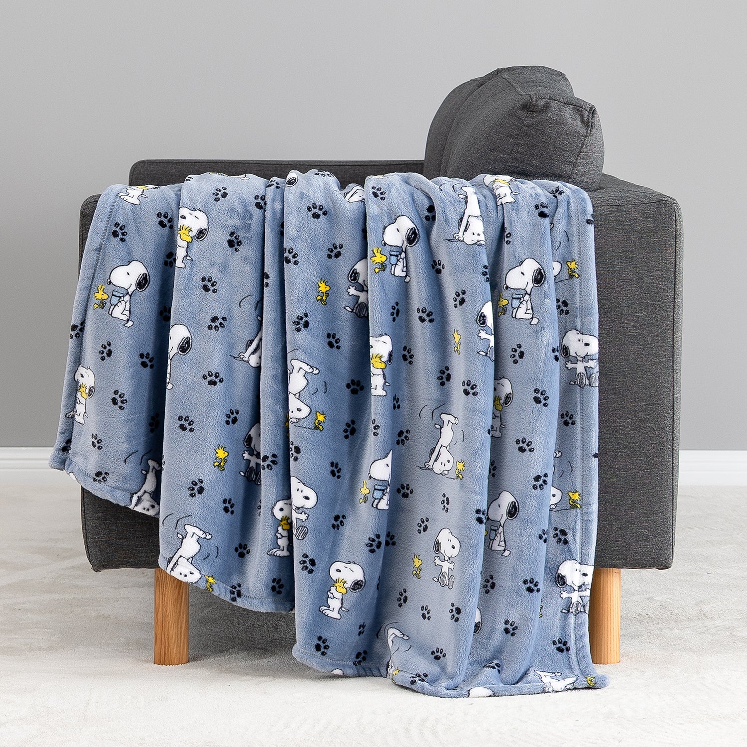 A light blue plush throw blanket with a repeating pattern of Snoopy, Woodstock, and paw prints, draped over a dark gray armchair.