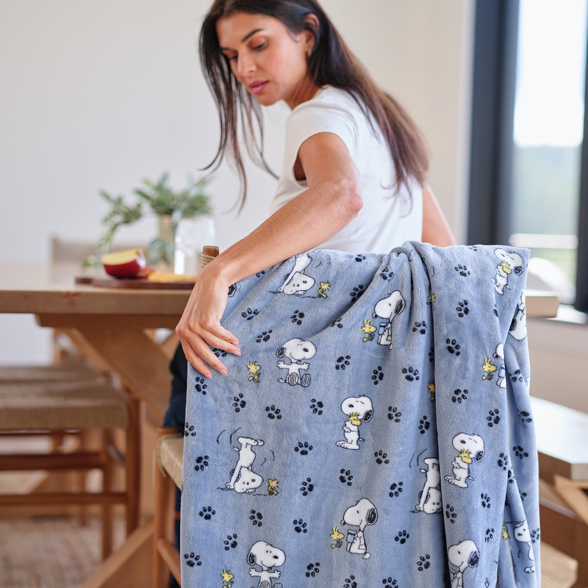 A woman sitting at a table touching a blye printed Peanuts throw blanket draped over her chair.